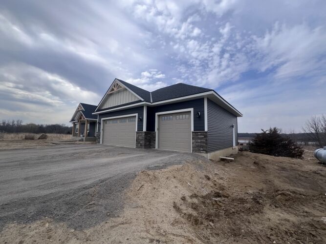 Modern house with gray siding and three-car garage on a gravel driveway, surrounded by bare soil and a cloudy sky.