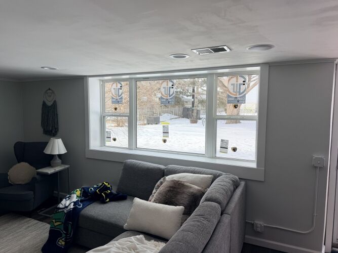 A living room with a gray sectional sofa faces three large windows. Snow is visible outside, and stickers are still on the new windowpanes.