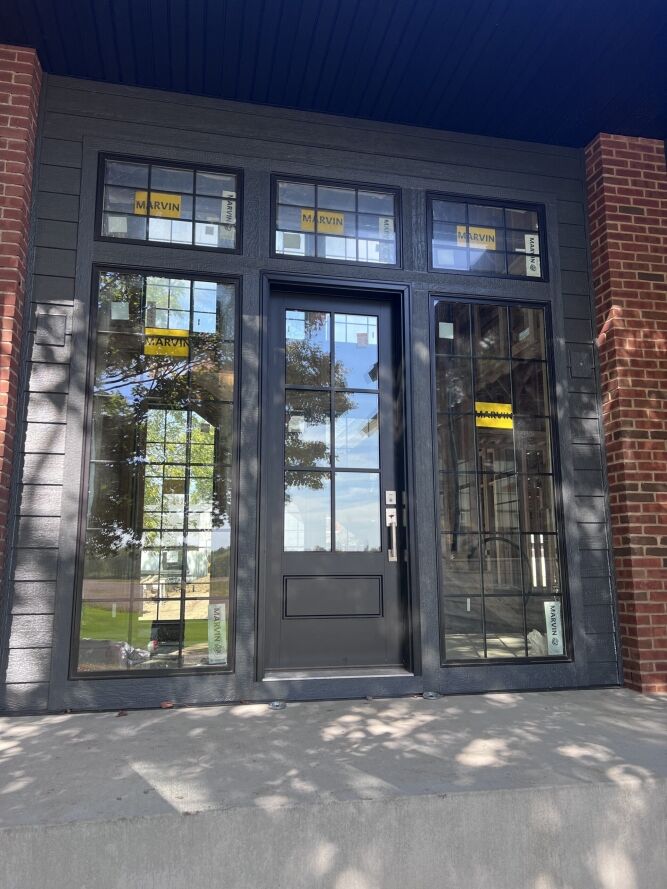 A modern black front door with glass panels is surrounded by tall side windows and transom windows, all displaying Marvin brand stickers.
