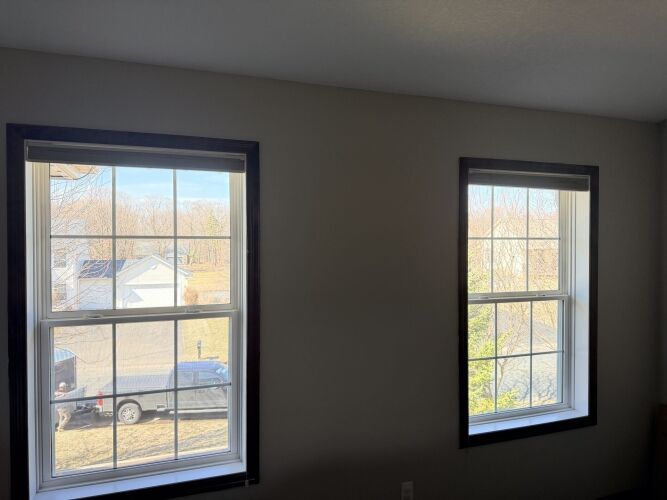 Two double-pane windows with dark trim are set in a beige wall, showing a view of a residential neighborhood with houses, trees, and a parked truck outside.