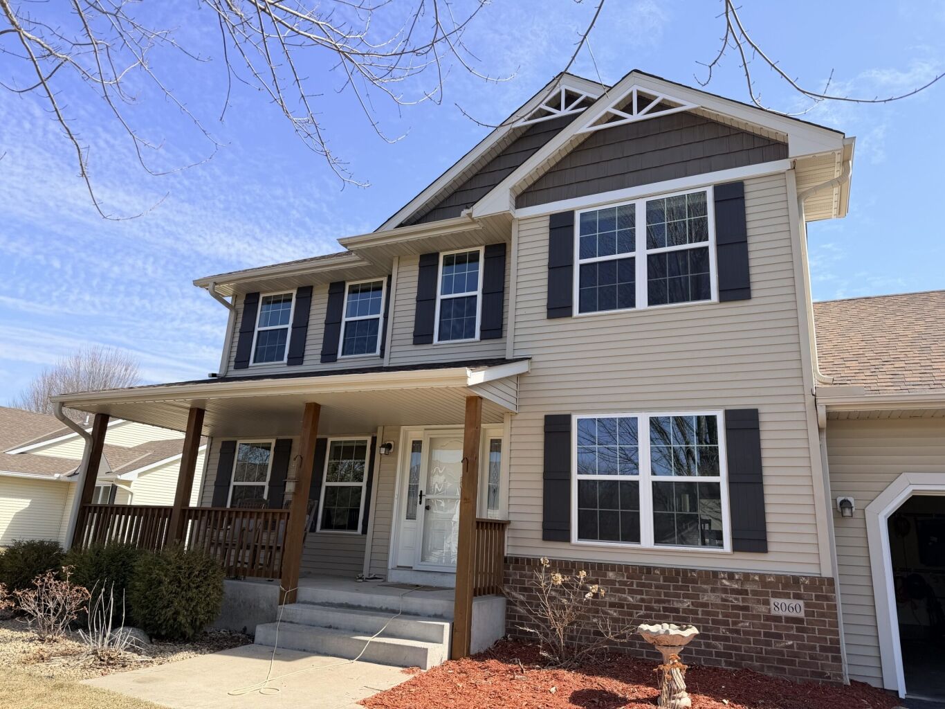 Two-story suburban house with beige siding, dark shutters, a covered front porch, and a brick foundation. The yard has mulch, bushes, and a concrete walkway.