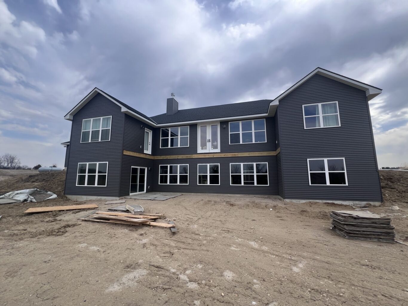 A large two-story house with dark siding and many windows, surrounded by dirt and construction materials, under a cloudy sky.