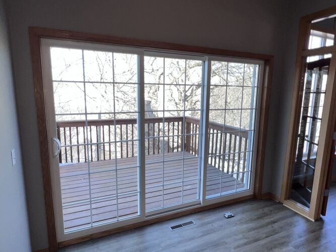 A glass sliding door with white grid frames opens to a wooden deck with railing, overlooking leafless trees. Indoors, there is wood-trim and gray flooring.