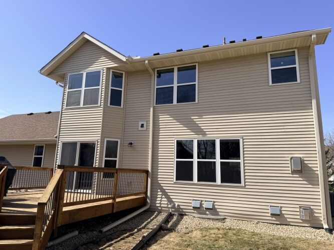 Two-story beige house with multiple windows, a wooden deck with railing, and a small gravel border along the foundation on a sunny day.
