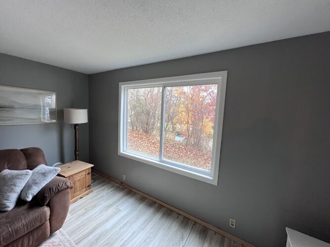 A living room with a brown couch, wooden side table, floor lamp, and a large window overlooking trees with autumn leaves.