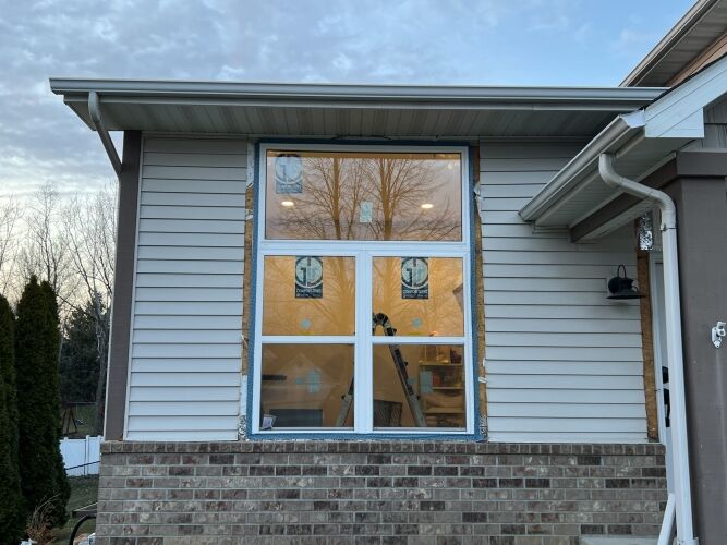 Large newly installed window with white frame on a house with beige siding and brick exterior; interior lights are on and construction materials visible inside.
