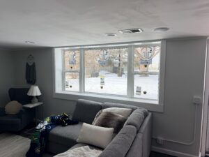 A living room with a gray sectional sofa faces three large windows. Snow is visible outside, and stickers are still on the new windowpanes.