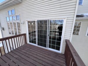 A wooden deck with a white sliding glass door and grid-pattern windows on the exterior of a house with beige siding.