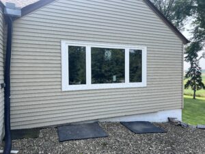 A beige-sided house with a large white-framed window, gravel ground below, two black mats, and lawn with trees visible in the background.