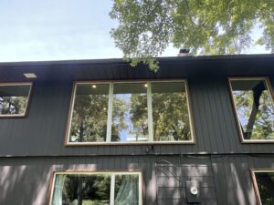 A dark gray two-story house with large windows reflecting trees, visible electrical meter, and overhead tree branches.