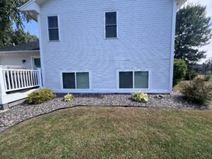 White house exterior with two basement windows, small shrubs, and plants along a rock mulch bed bordered by lawn.