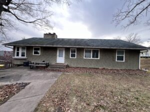 Single-story house with a green shingle roof, wood siding, several windows, a white door, and a concrete patio with a picnic table; lawn and bare trees surround the home.