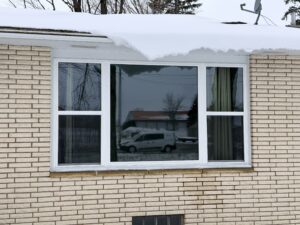 A snow-covered roof overhangs a white-framed window on a brick house, with a white van and trees reflected in the glass.