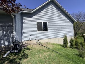 The side of a light blue house with a single window, garden hose reel, gas meter, three small evergreen shrubs, and a grassy yard under a clear blue sky.