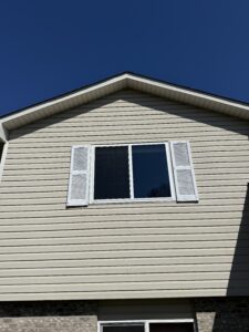 The upper exterior of a house with beige siding, a double window with white shutters, and a clear blue sky in the background.