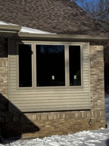 A house exterior showing three large windows on a beige, brick and siding wall, with snow on the ground and roof.