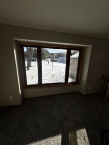A large window overlooks a snowy yard with trees, a streetlamp, and a house in the background. Sunlight casts shadows inside the carpeted room.