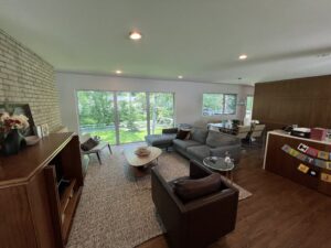 Modern living room with large windows overlooking a green backyard, featuring a sectional sofa, armchair, coffee table, and birthday decorations on a cabinet.