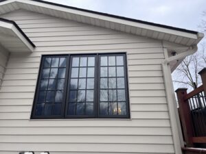 A beige house exterior with black-trimmed grid windows reflecting trees and sky, next to a white gutter and a wooden stair railing.