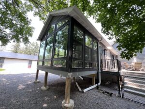 A modern glass sunroom with large windows is elevated on wooden posts, attached to a house, surrounded by trees and gravel ground.