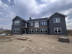 A large two-story house with dark siding and many windows, surrounded by dirt and construction materials, under a cloudy sky.