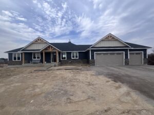 Single-story modern house with dark blue siding, stone accents, and a three-car garage, set on a dirt lot under a cloudy sky.