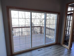 A glass sliding door with white grid frames opens to a wooden deck with railing, overlooking leafless trees. Indoors, there is wood-trim and gray flooring.