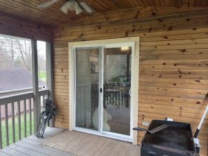 A covered wooden porch with folding chairs, a grill, a doormat, and a glass sliding door leading to the interior of the house.