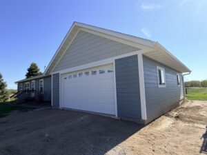Single-story house with blue-gray siding, white trim, and an attached two-car garage on a gravel driveway under a clear blue sky.