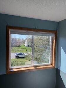 A window with a wooden frame overlooks a suburban street with houses, trees, and a parked white SUV on a sunny day.