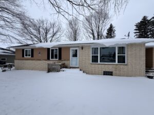 A single-story house with beige siding and brick exterior, covered in snow, surrounded by bare trees and an overcast sky.