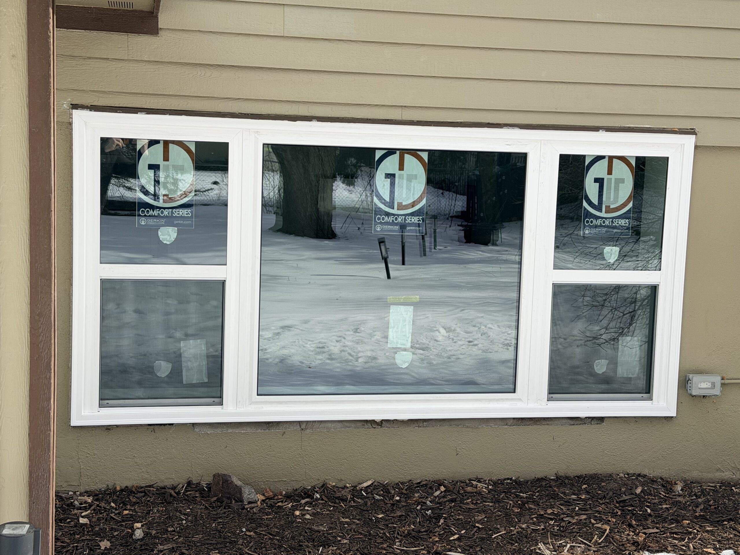 Three-panel window with white framing installed in a beige wall, with two stickers labeled Comfort Series on the glass. Snow and trees are visible outside.