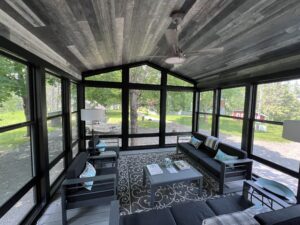A screened-in porch with dark furniture, a patterned rug, a ceiling fan, and large windows providing views of a green yard and trees outside.